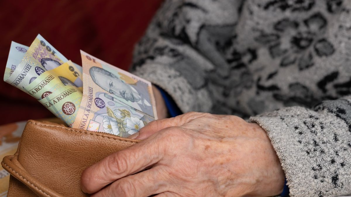 O bătrână cu pulover pe ea scoate dintr-un portofel maro, pe care îl ține cu o mână, câteva bancnote românești