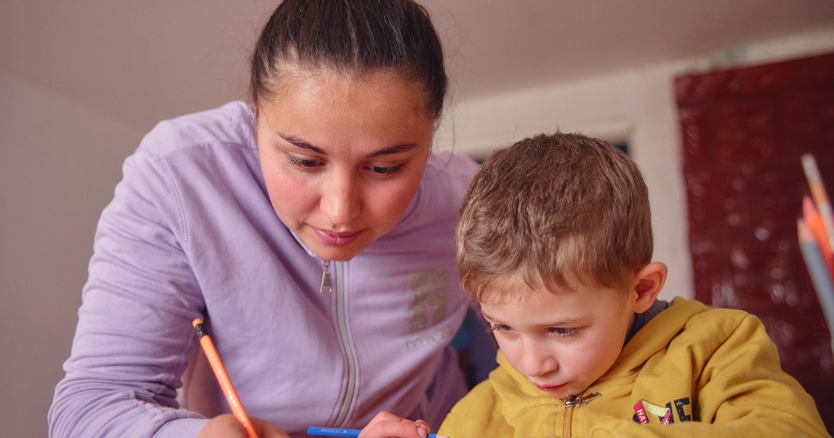 O mamă și fiul ei care sunt aplecați peste o masă și lucrează împreună la lecții. Amândoi folosesc creioane colorate, mama scriind pe hârtie în timp ce îl ghidează pe copil.