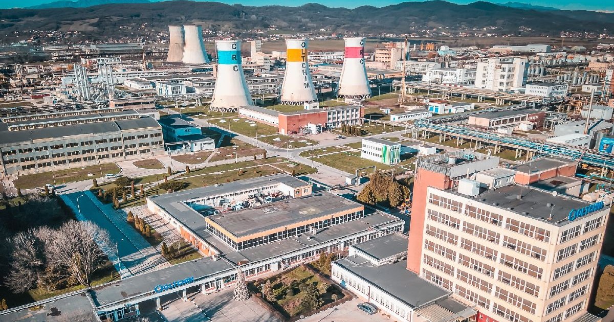 Imaginea prezintă o vedere aeriană amplă a unei platforme industriale. În plan îndepărtat, se remarcă mai multe turnuri de răcire masive. În prim-plan și în partea dreaptă se află clădiri de birouri.