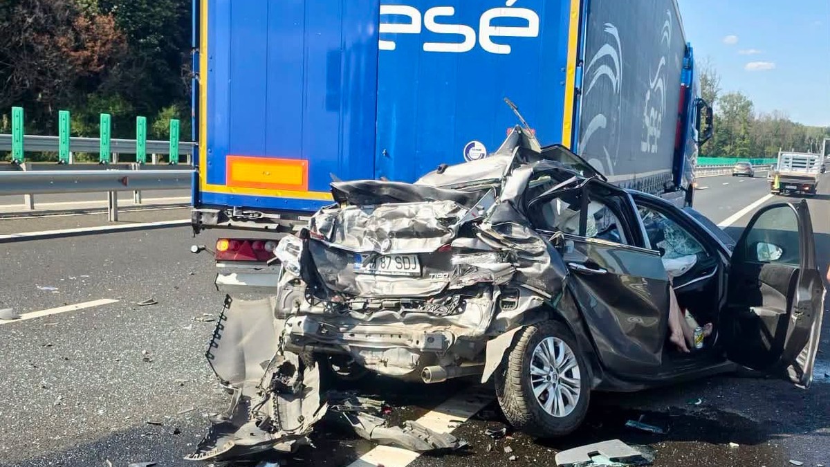 În imagine se vede un autoturism grav avariat, prins în partea din spate sub remorca unui camion. Caroseria este puternic deformată, iar resturi ale mașinii sunt împrăștiate pe carosabil.