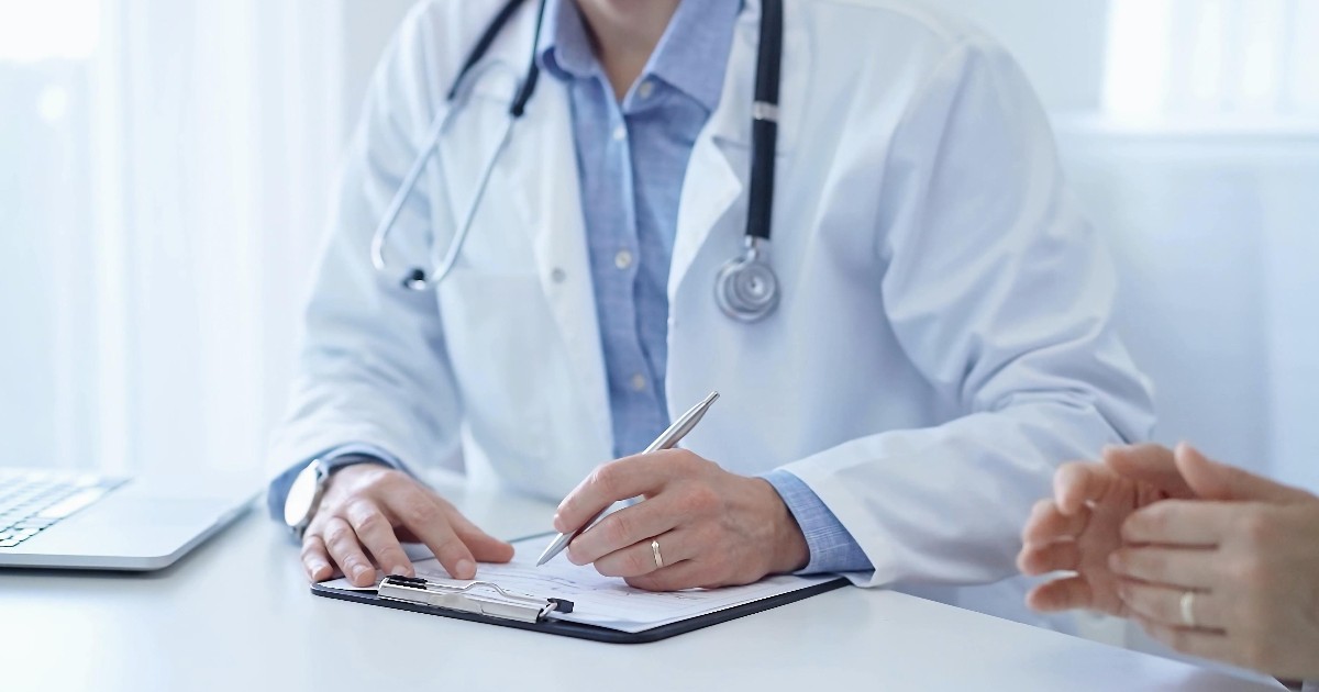 Un medic care stă la birou, în cabinetul său, poartă un halat alb și scrie o recomandare unei paciente
