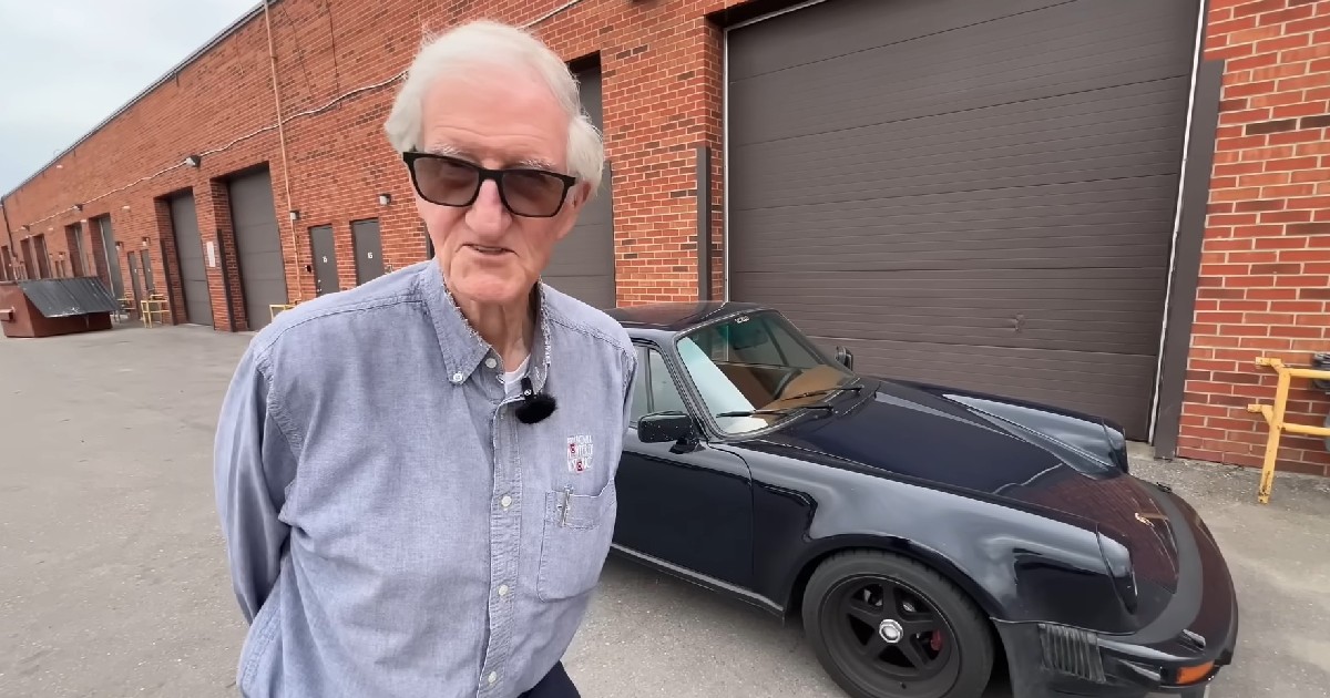 Imaginea prezintă un bărbat cu ochelari și cămașă albastră stând în fața unui automobil Porsche clasic de culoare închisă, parcat lângă o clădire din cărămidă roșie.