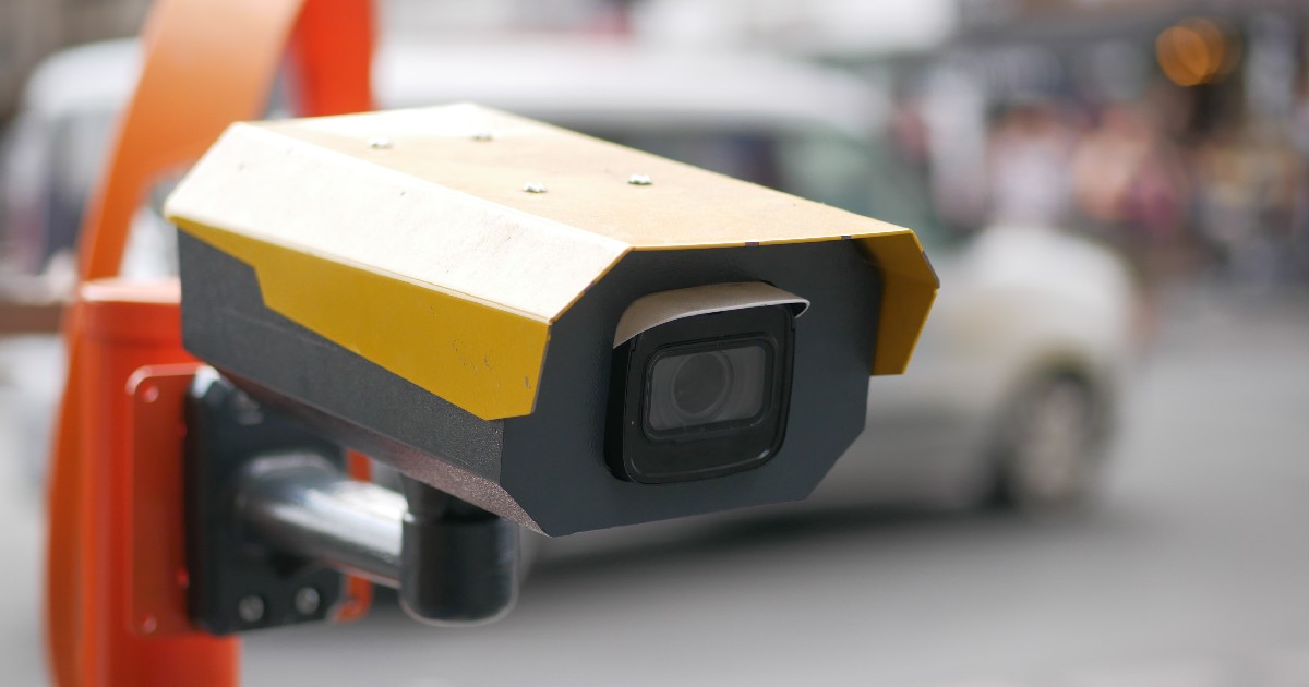 Un radar cu o carcasă galbenă este montat pe un stâlp portocaliu într-o zonă urbană aglomerată, având pe fundal traficul estompat al orașului.