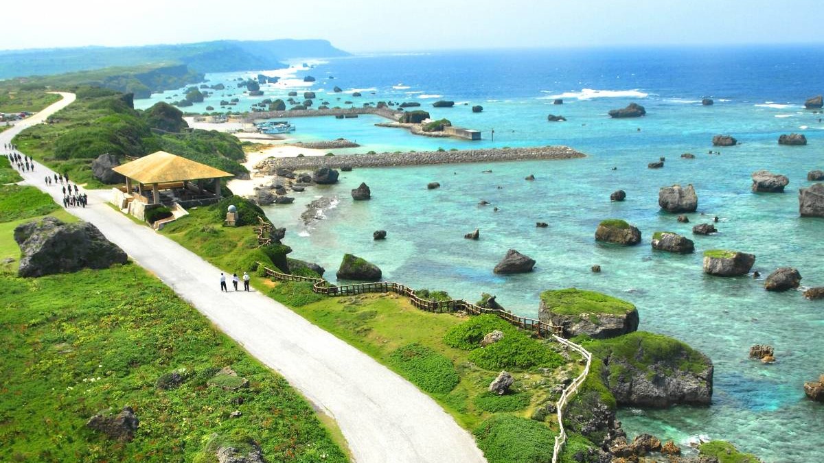 Imaginea arată o coastă din Okinawa, Japonia, cu apă turcoaz plină de stânci ieșite la suprafață. În stânga se vede un drum alb pe care merg grupuri de oameni, iar lângă el un foișor cu acoperiș galben. Țărmul este verde și stâncos, iar în depărtare se întinde marea albastră până la orizont.