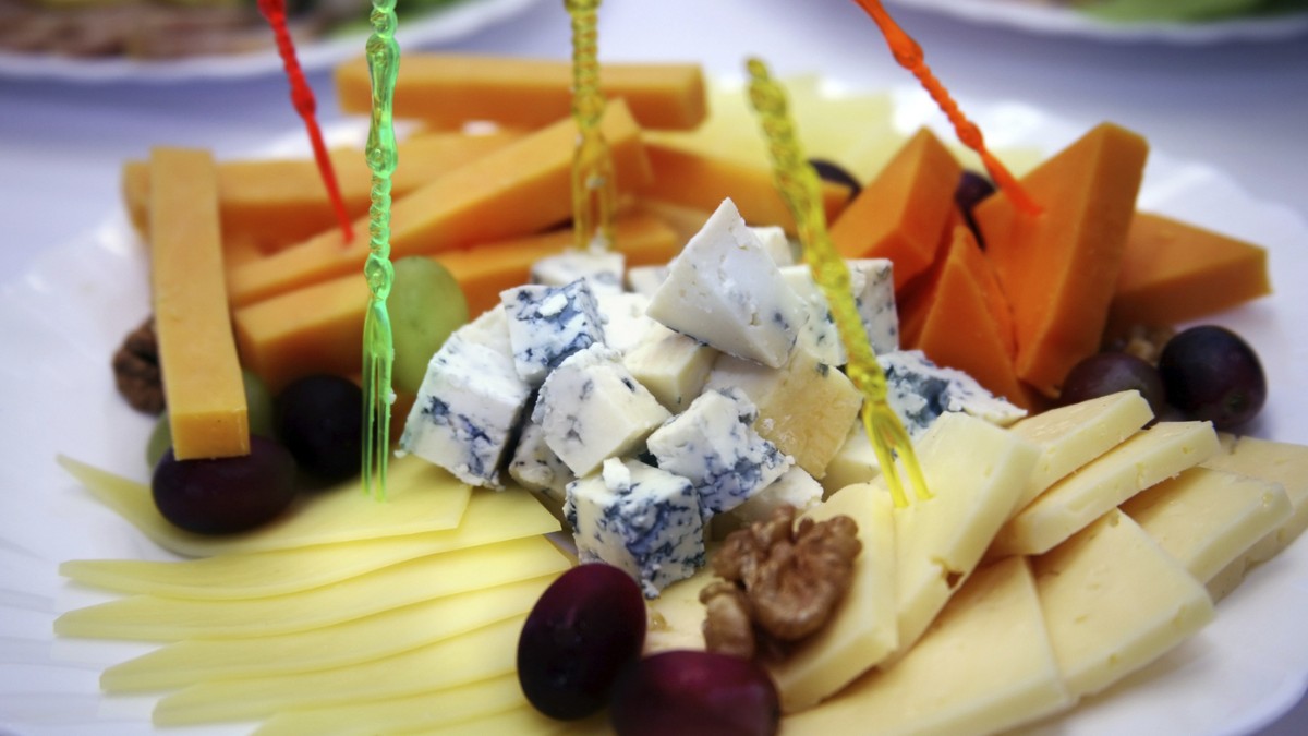 Imaginea prezintă un platou variat de brânzeturi, aranjate elegant pe o farfurie albă. Se pot observa mai multe tipuri de brânză – unele galbene, tăiate în felii sau bastonașe, și altele albe, cu mucegai albastru – alături de struguri, nuci și măsline, toate fixate cu bețișoare decorative colorate.