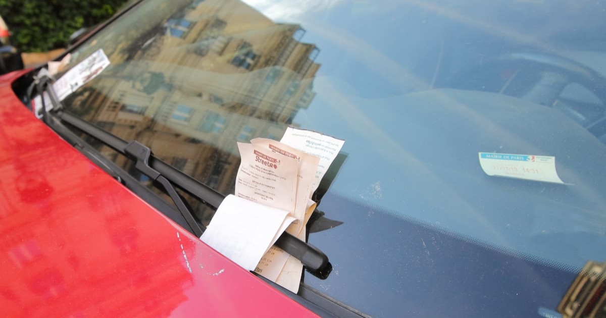 Imaginea prezintă mai multe amenzi de parcare puse sub ștergătorul uneii mașini roșii