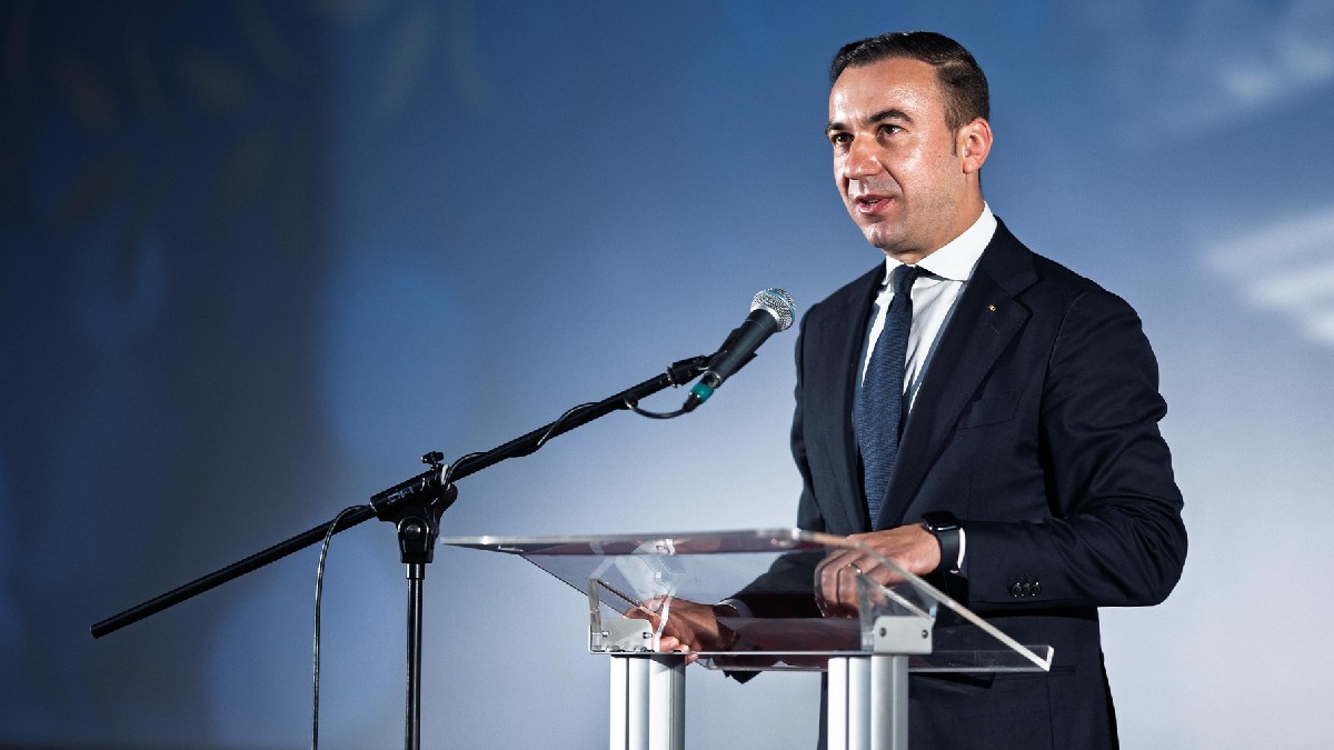 Bogdan Ivan, îmbrăcat într-un costum negru cu cămașă albă cu o cravată verde, în timpul unor declarații de presă, stând la un pupitru din plastic transparent, cu mâinile așezate pe el, vorbind la un microfon. Fundalul este de o culoare albastru deschis