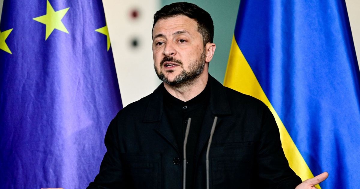 Volodimir Zelenski, îmbrăcat într-o cămașă neagră, dă explicații cu mâinile larg deschise în timpul unei conferințe de presă, având în spate steagurile Ucrainei și al Uniunii Europene