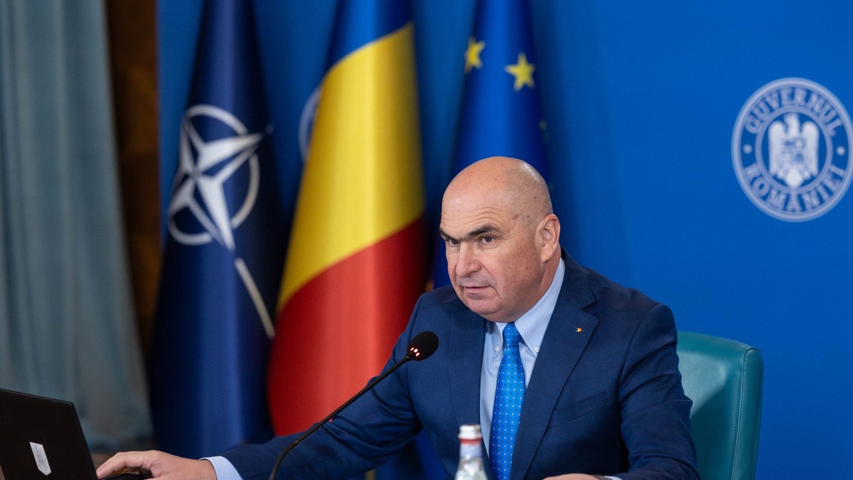 Ilie Boloja, premierul României, așezat la masă, îmbrăcat în costum bleu marin si cu cravata albastra. In spate, pe fundal albastru, se vâd steagul NATO, cel al României și steagul UE