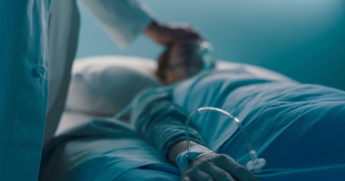Pacientă spitalizată întinsă într-un pat de spital noaptea, un medic o îngrijește, mâna în prim-plan.