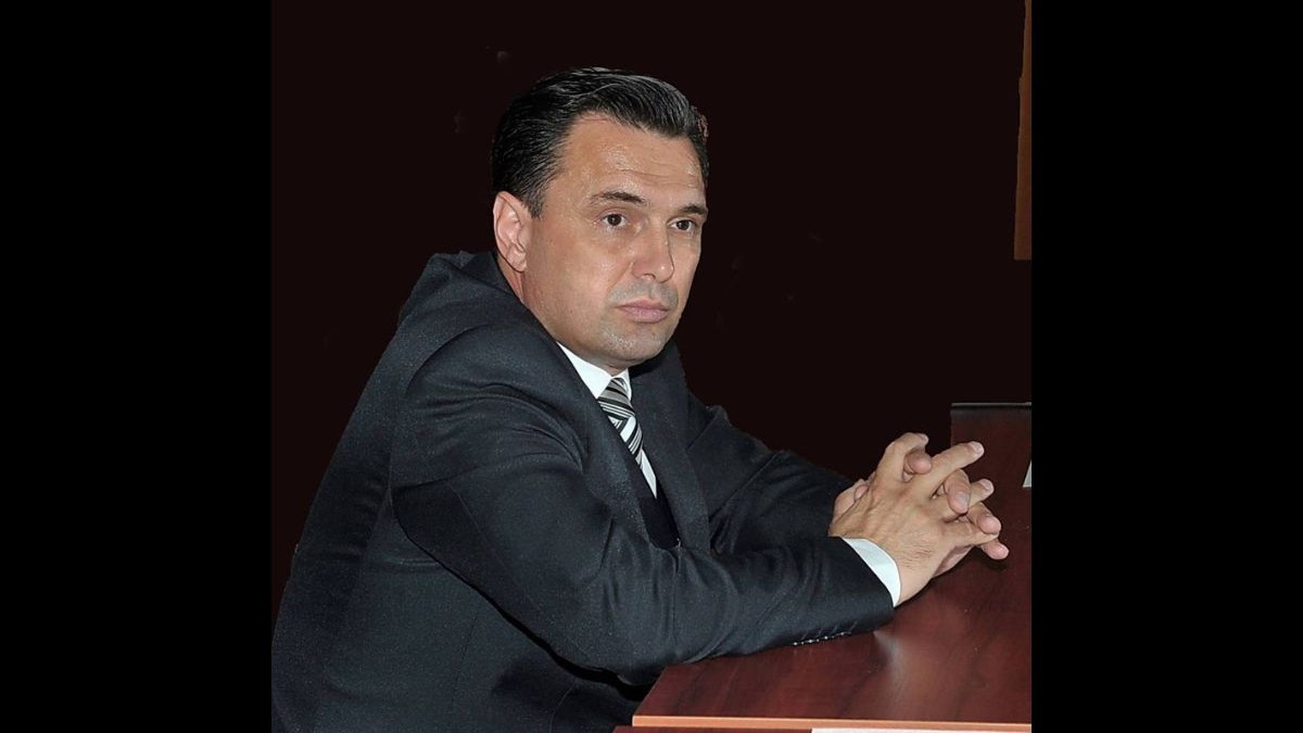 Liviu Negoiță, primarul municipiului Cernavodă, cu o expresie serioasă, stând cu mâinile împreunate pe masă, îmbrăcat într-un costum negru, o cămașă neagră și o cravată în dungi