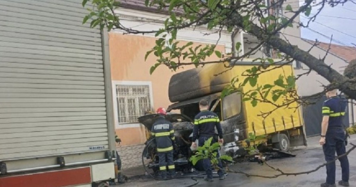 În imagine este ilustrată o camionetă galbenă, care a ars. De asemenea, se observă și pompieri și polițiști în apropierea acesteia