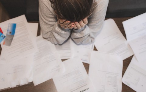 O persoană care își ține capul în mâini deasupra unei mese pline de facturi, documente împrăștiate și carduri bancare, sugerând o stare de stres sau epuizare financiară