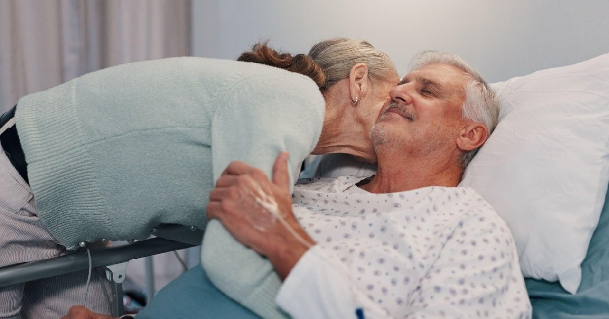 Femeie in varsta imbratisand un barbat cu parul alb, in pijama alba, pe un pat de spital