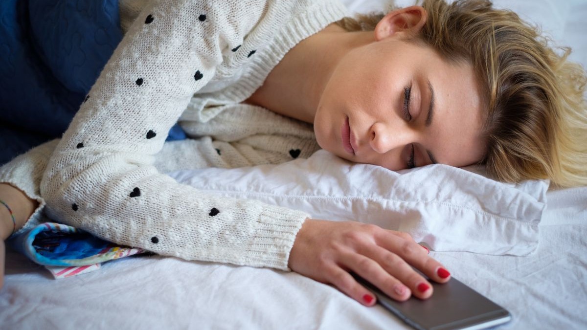 O tânără blondă, cu un pulover alb cu inimioare negre, doarme pe un pat pe care este un cearșaf alb și ține o mână pe telefonul mobil