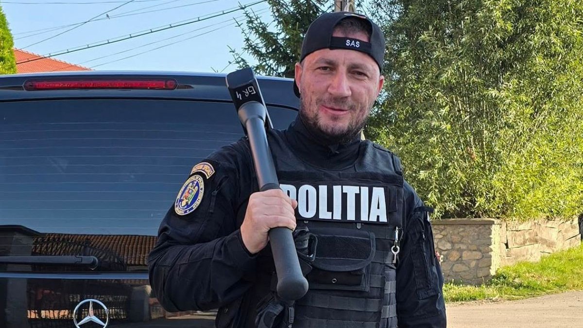 Marian Godină, într-o uniformă neagră de Poliție, cu șapca întoarsă la spate pe care scrie SAS și cu un dispozitiv de deschis uși într-o mână, pe umăr