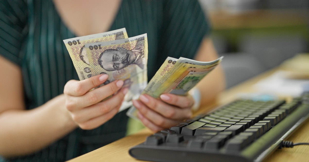 O femeie cu rochie verde care stă la birou și numără mai multe hârtii de 100 de lai