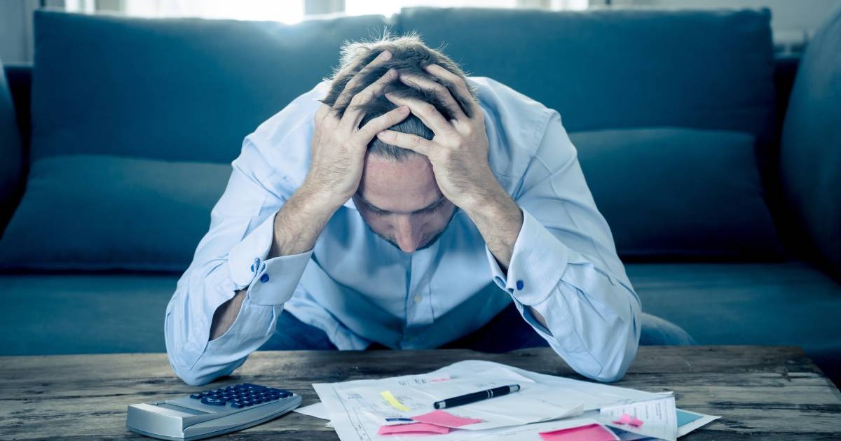 Barbat stresat, sta pe canapea cu mainile la cap si se uita la facturi rate la bancă, puse pe o masa