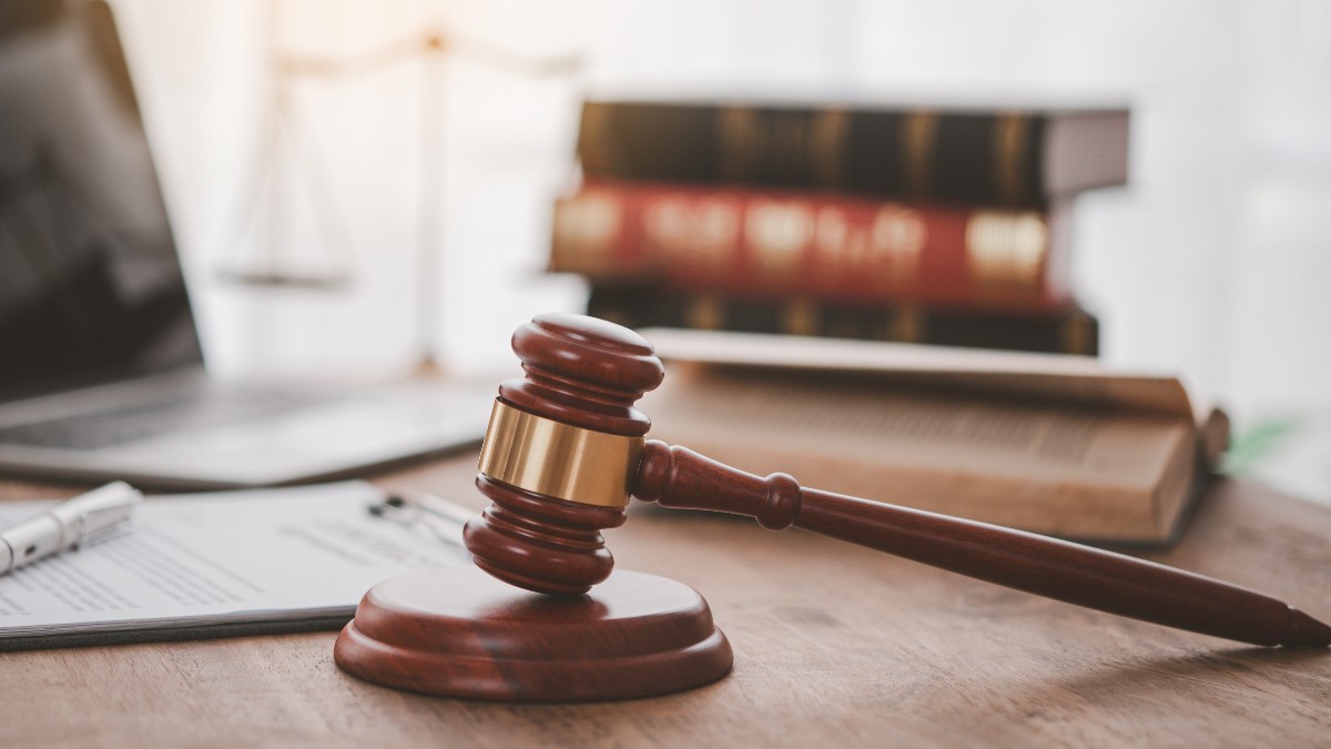 Ciocanul unui judecător pe o masă de lemn, alături de cărți de drept, o balanță a justiției și un laptop.