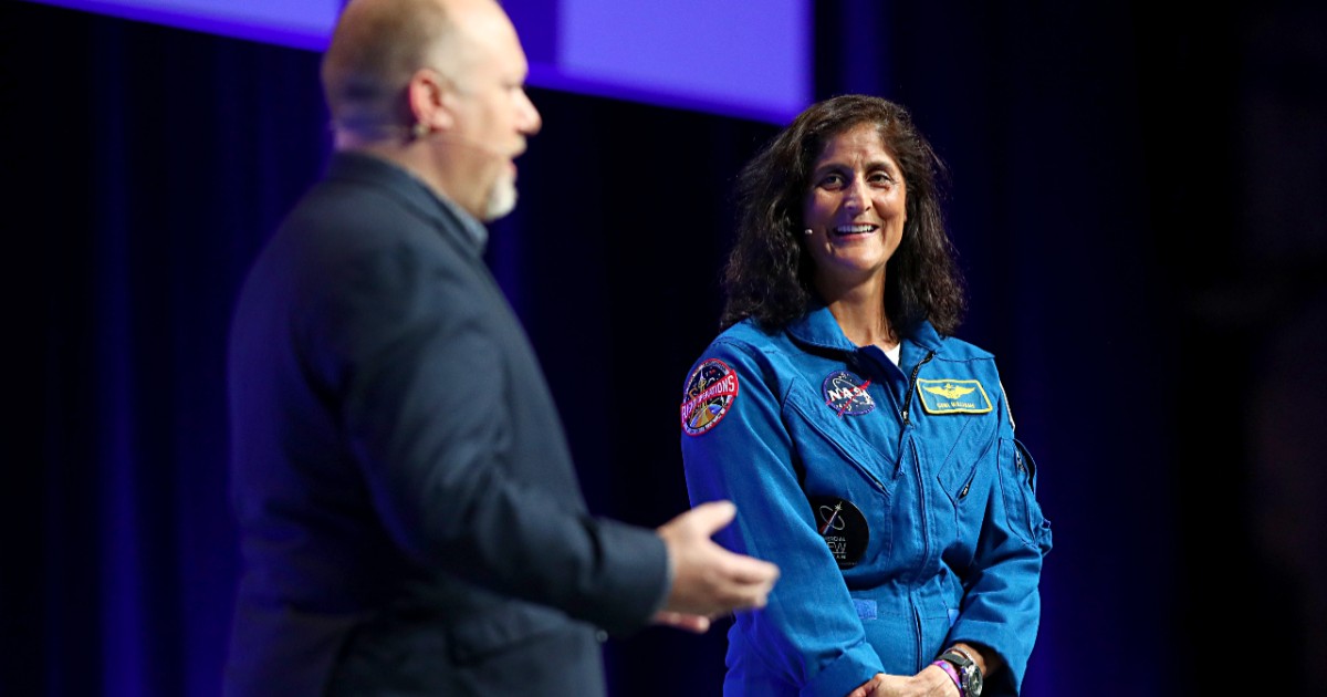 Sunita Williams în costum albastru de astronaut, pe o scenă, lângă un bărbat în costum negru.
