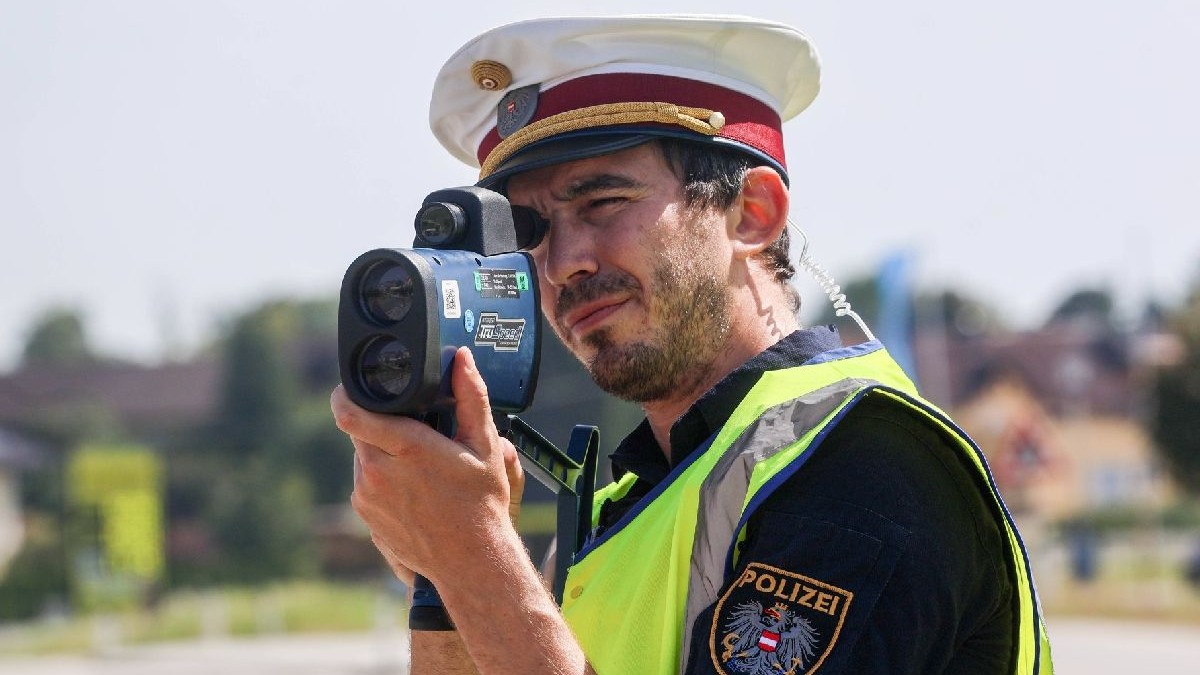 Un bărbat cu barbă, îmbrăcat în uniformă de polițist și cu o șapcă albă cu cozoroc ține un parat radar la ochi