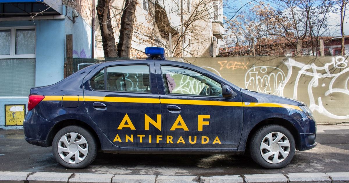 O mașină marca Dacia Logan, cu logo-ul ANAF Antifraudă, de culoare albastră, cu text galben, parcată pe trotuar, lângă un gard de beton desenat cu graffiti
