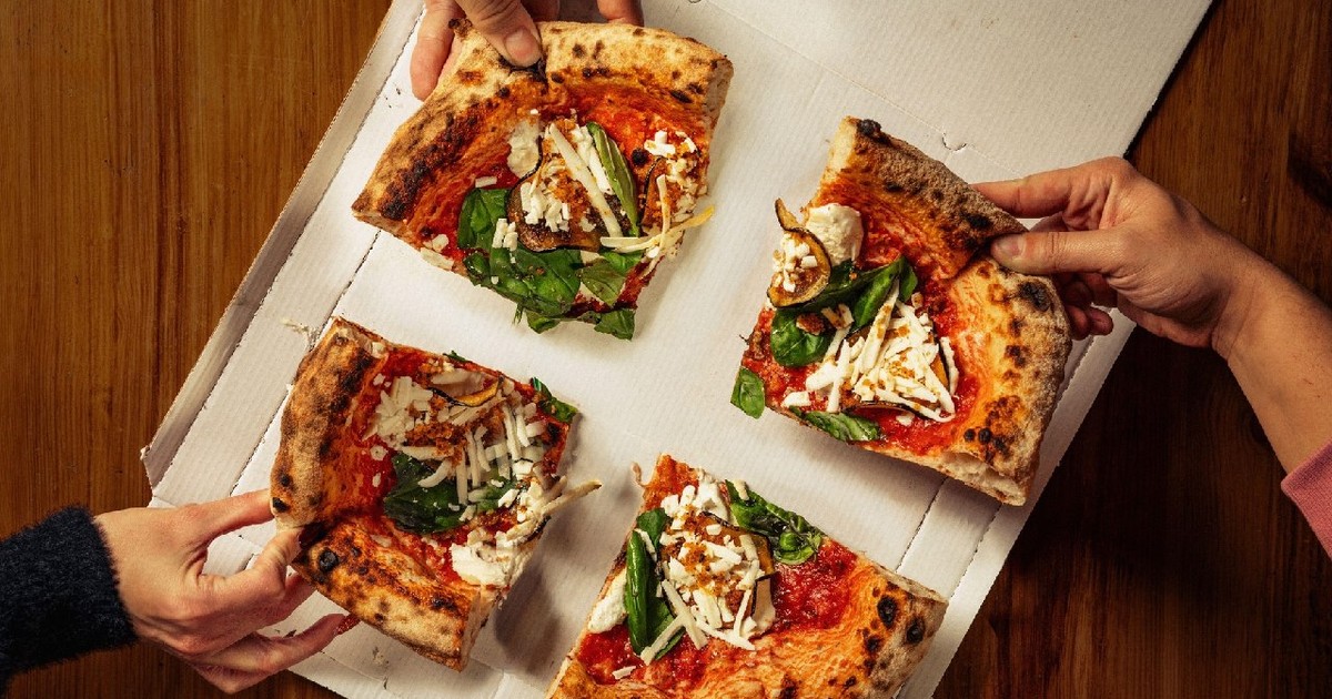 Patru mâini care iau felii de pizza cu brânză, busuioc și sos de roșii dintr-o cutie de pizza așezată pe o masă de lemn.