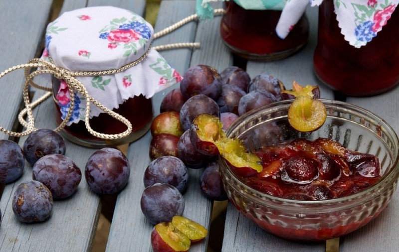 Un bol cu dulceață de prune, prune întregi și borcane de gem pe o masă de lemn