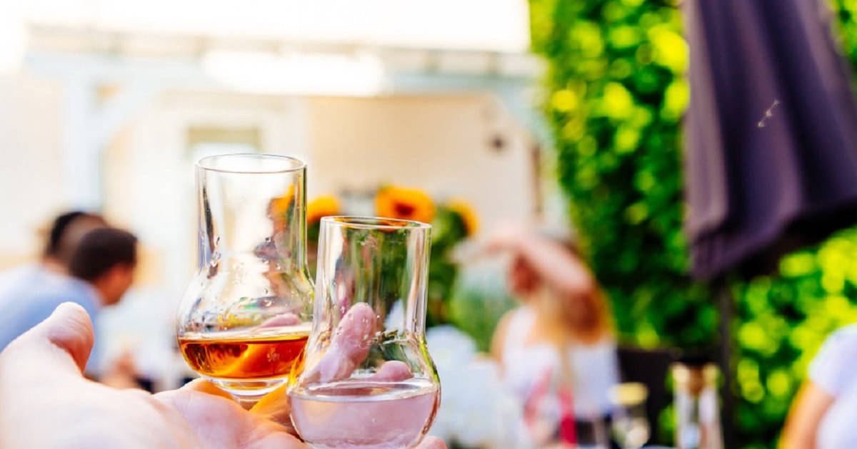 De unde vine tradiția digestivelor – cât e mit și cât e realitate - Imagine cu o mână bărbătească ţinând două pahare mici cu grappa, unul cu lichid de culoare castanie şi unul incolor.
