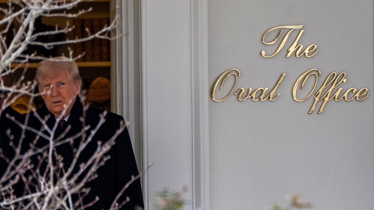 Donald Trump, îmbrăcat în pardesiu negru, în spatele unui copac desfrunzit, iar în dtânga sa scrie The Oval Office