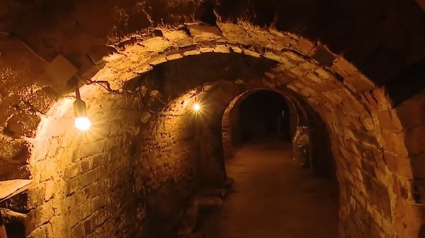 Interiorul unui tunel sau al unui beci subteran vechi, zidit din cărămidă roșie cu tavane boltite, iluminat de becuri.
