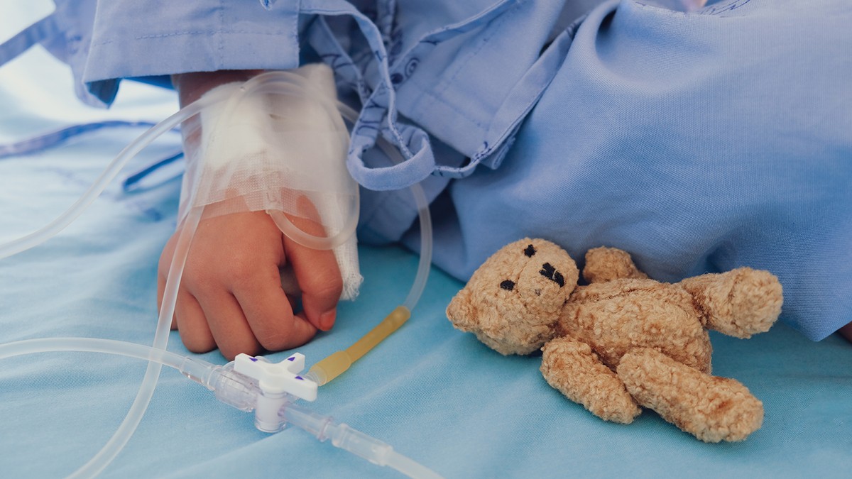 Un copil bolnav pe patul de spital alaturi de un ursulet de plus