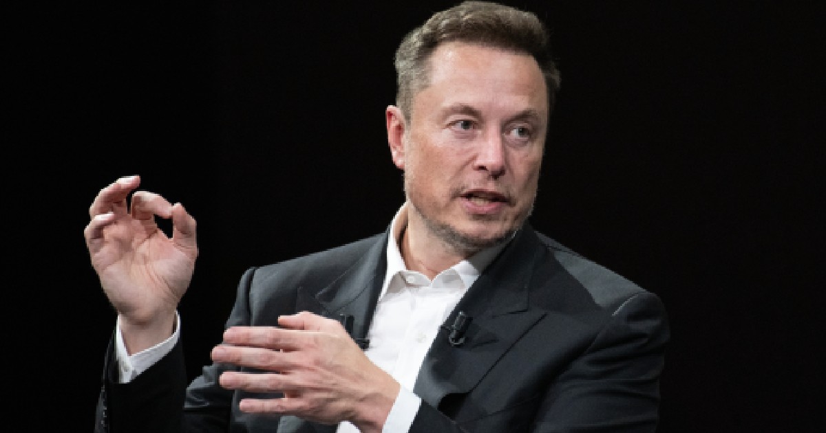 Imaginea îl înfățișează pe Elon Musk purtând un costum negru și o cămașă albă, în timp ce gesticulează cu mâinile la o prezentare sau interviu, pe un fundal negru.