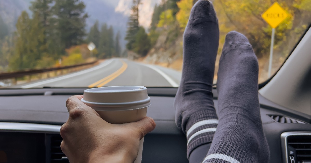 Femeie pe scaunul pasagerului, ținându-și picioarele și o ceașcă de cafea pe bord, în timp ce călătorește prin munți, toamna