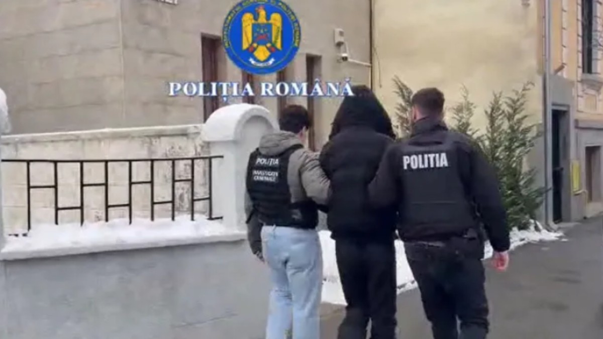 Un tânăr îmbrăcat cu pantaloni și geacă neagră, ținut de doi polițiști, pe drum.