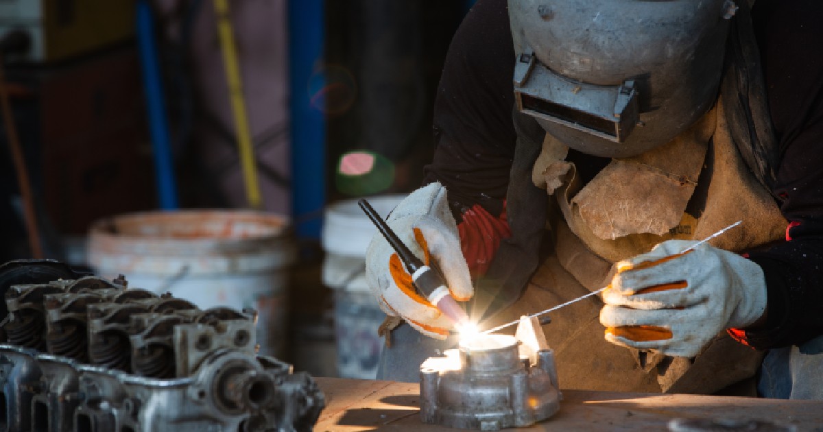 Imaginea prezintă un lucrător echipat cu mască de protecție și mănuși care sudează cu precizie o piesă metalică.
