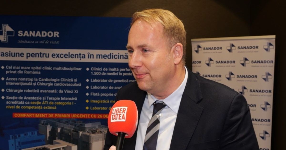Dr. Victor Costache, îmbrăcat cu costum negru, cămașă albă și cravată neagră, cu microfonul Libertatea în față și un panou cu Sanador pe fundal