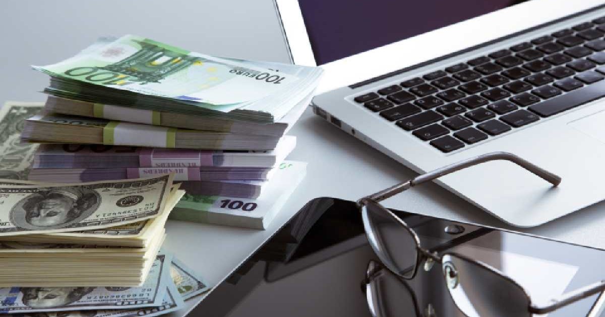 Pe o masă se află un laptop, un teanc de bani (dolari), o tabletă și o pereche de ochelari