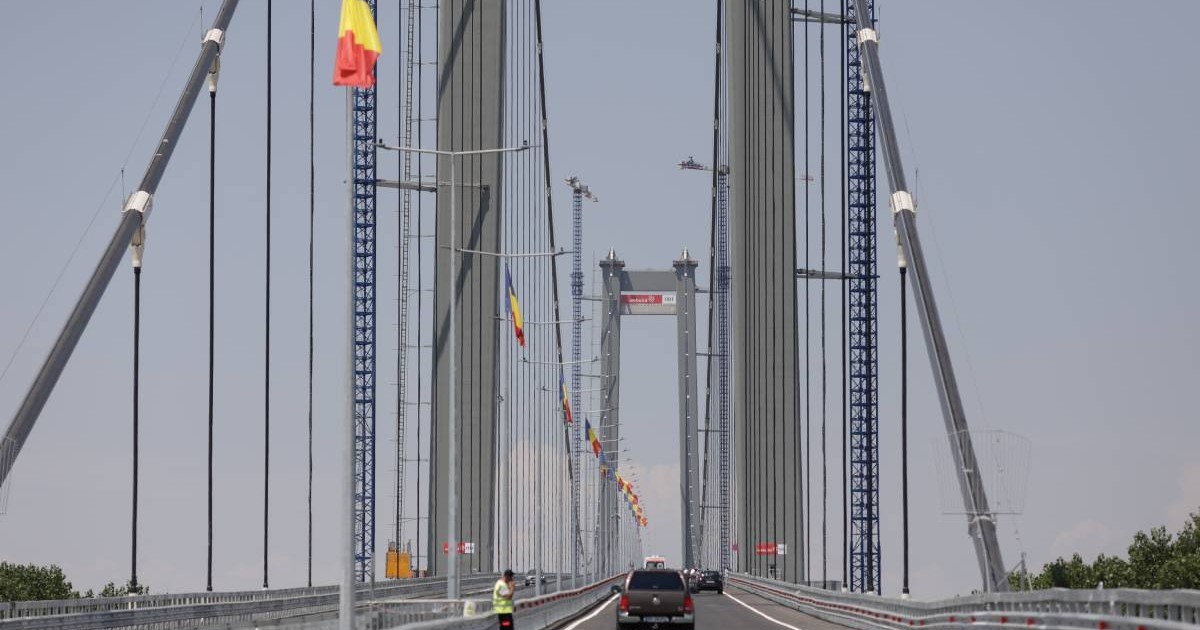 podul suspendat peste Dunăre de la Brăila, cu mașini care circula pe drum și stegulețe tricolore arborate de-a lungul cablurilor de susținere, fără trafic intens