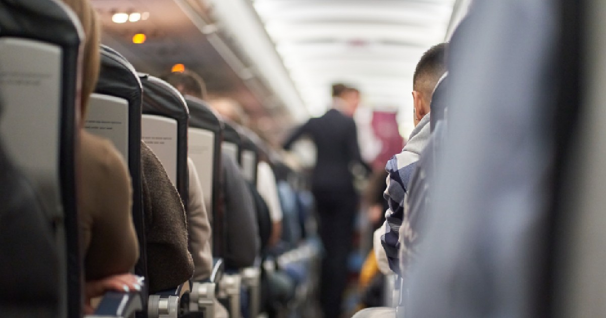 Mai mulți pasageri stau pe scaunele din avion. În fața lor se află un însoțitor de zbor