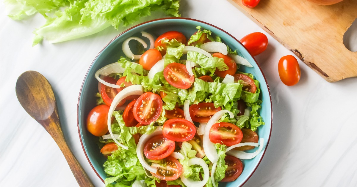 Imaginea prezintă o salată proaspătă, colorată, alcătuită din frunze de salată verde, roșii cherry și felii subțiri de ceapă. Preparatul este așezat într-un bol, lângă ingrediente proaspete și o lingură de lemn, sugerând un stil de viață sănătos.