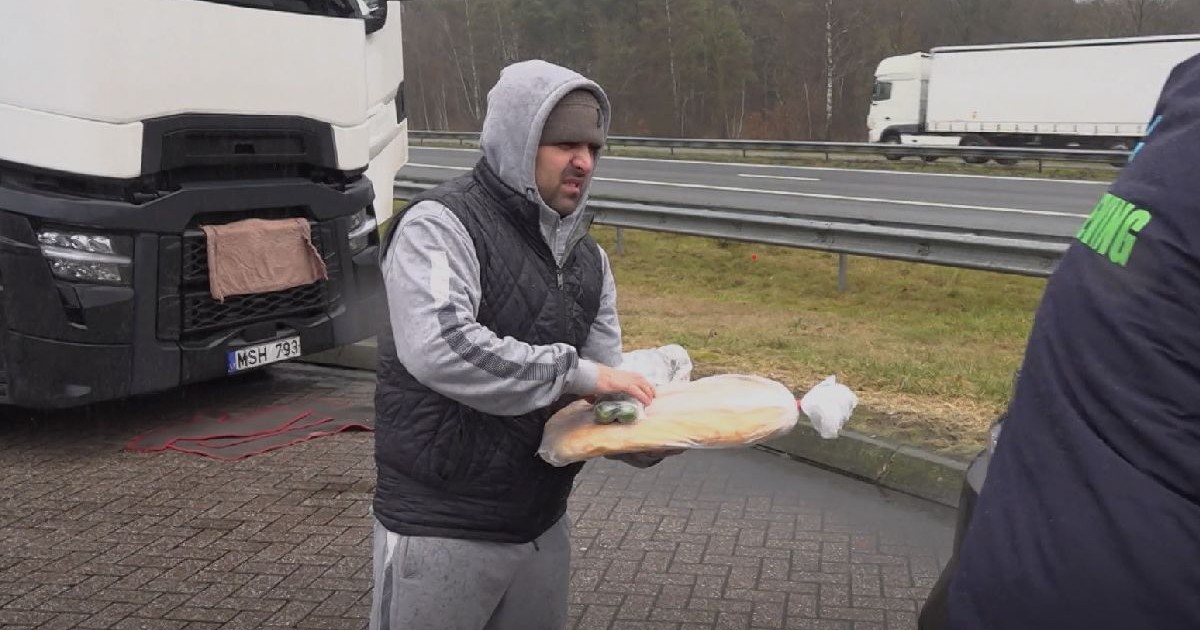 Un bărbat îmbrăcat într-un hanorac gri cu glugă și vesta neagră ține în mâini o pâine, într-o parcare, lângă un camion alb.