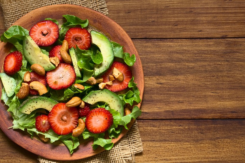 55971795 - strawberry, avocado, lettuce salad with cashew nuts on plate, photographed overhead on dark wood with natural light