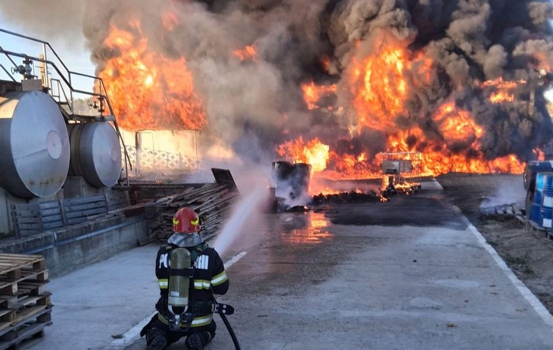 În curtea unei hale industriale, un pompier echipant cu aparat de respirat stă în genunchi pe o platformă betonată și direcționează un jet de apă spre un incendiu violent, sutești, valcea, 3 octombrie 2026