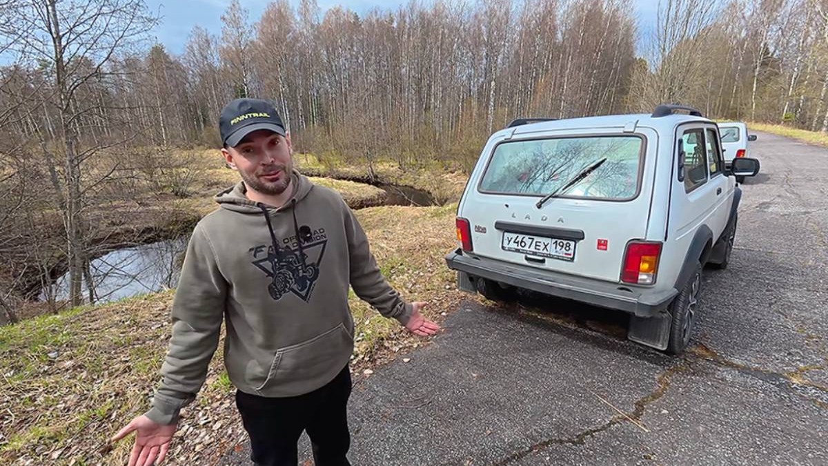 Youtuber rus stând lângă o mașină Lada Niva, parcată pe marginea unui drum de pădure.