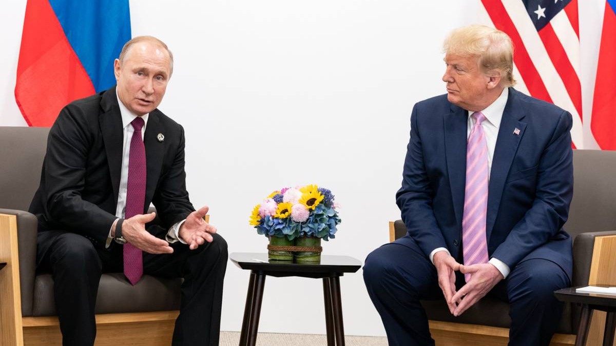 intalnire dintre vladimir putin si donald trump, cei doi sunt asezati pe cate un scaun. in partea din stanga este vladimir putin care gesticulează cu mâinile, în timp ce in dreapta este donald trump care îl ascultă atent, având o expresie serioasă. pe fundal sunt steagurile Rusiei și SUA
