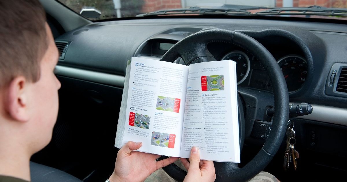 Un tânăr cu părul scurt citește dintr-o carte cu teste auto în timp ce se află la volan, pe scaunul din dreapta, într-o mașină parcată