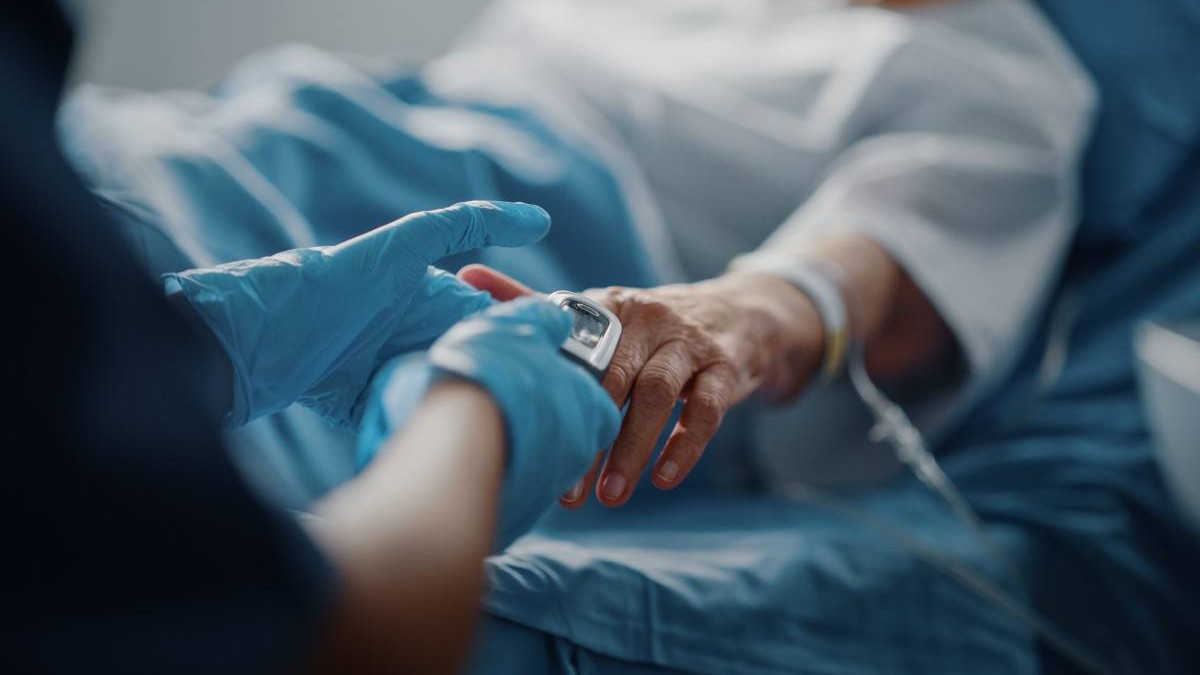 O femeie în vârstă pe patul de spital, după o intervenție, are puls oximetru montat la mână, iar o asistentă o îngrijește.