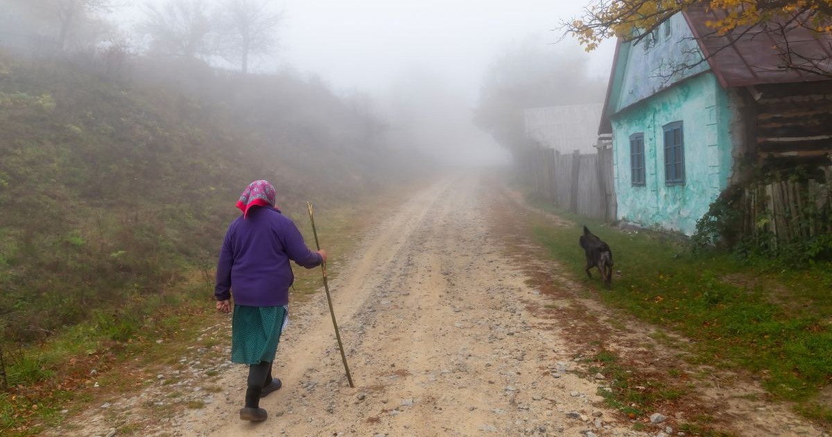 Femeie bătrână, îmbrăcată cu o fustă verde și un pulover albastru, cu un batic roșu pe cap, pe uliță cu un băț în mână, lângă o casă vopsită verde