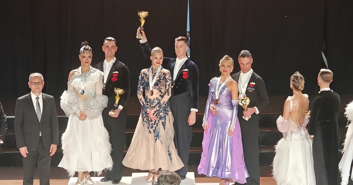 O fotografie de la ceremonia de premiere a unui concurs de dans sportiv, unde trei perechi de finaliști, îmbrăcate elegant în costume de competiție și rochii spectaculoase, stau pe podium alături de un oficial, în timp ce perechea câștigătoare ridică trofeul, totul desfășurându-se pe o scenă cu parchet și un fundal negru, în fața unor cameramani.